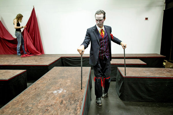 A man in a 3 piece suit and white stage facial makeup peruses a theater set with two walking sticks.