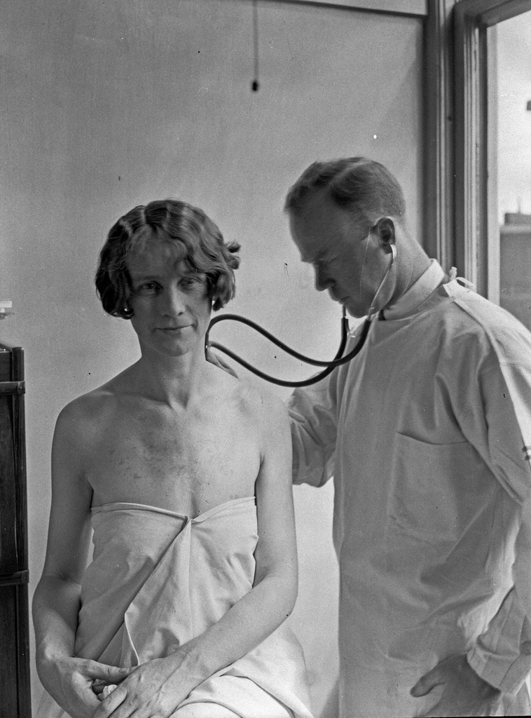 A back and white photo of a woman patient and a male doctor, likely from the first half of the 20th century,. The woman sits with a sheet wrapped around her torso, and the dotor listens to her back with a stethoscope.