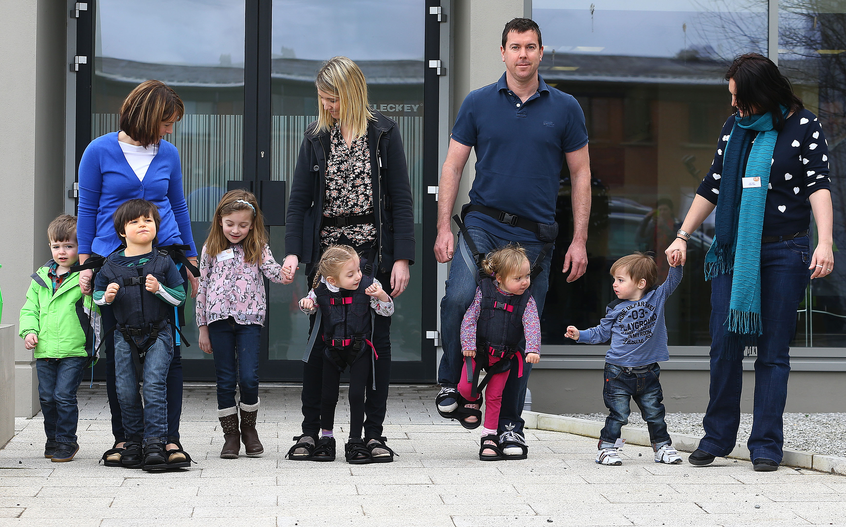 Several adults "wear" their young toddlers in the Firely Upsee baby carriers, but inverted: the child is harnessed to an adult's legs and feet and strapped securely via the shoulders to the adult's waist, allowing simultaneous walking for both caregiver and young user.