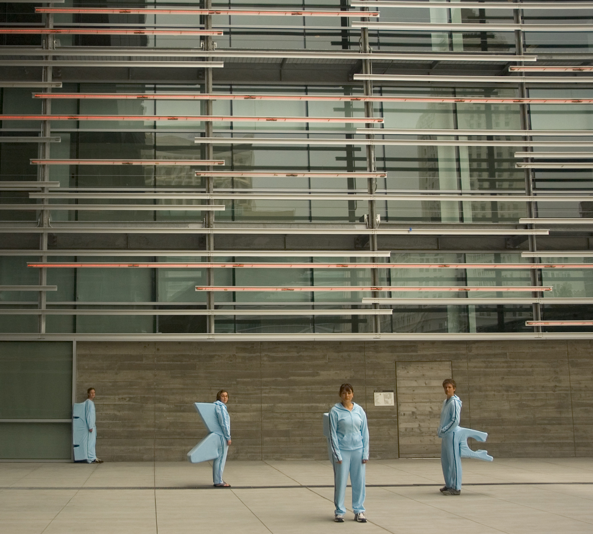 Four people wear pale blue, soft knit jogging suits, with soft geometric forms attached to their backs. Each form fits precisely over a designated architectural site, making it possible for a person to perch or sit at that site...