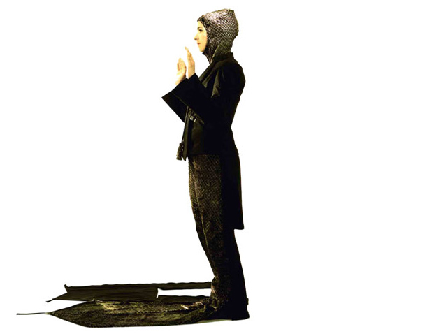 a woman wears an all-black suit, with blazer and coat, complete with unfolded prayer rug from the leg section of fabric, unzipped and spread on the floor, and a head scarf pulled from the back of the jacket.