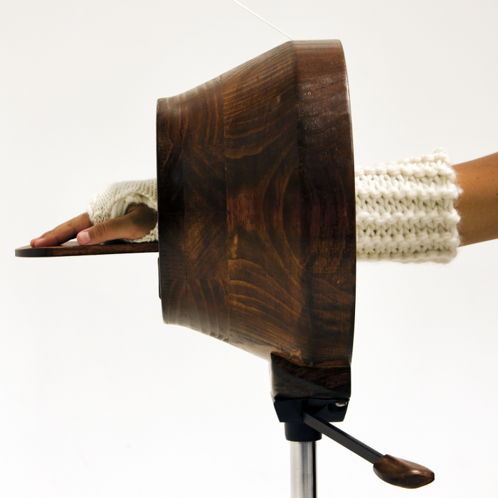 a round, hollow wooden hand-cranked knitting machine creates a wool cast for a human arm at the limb passes through it.