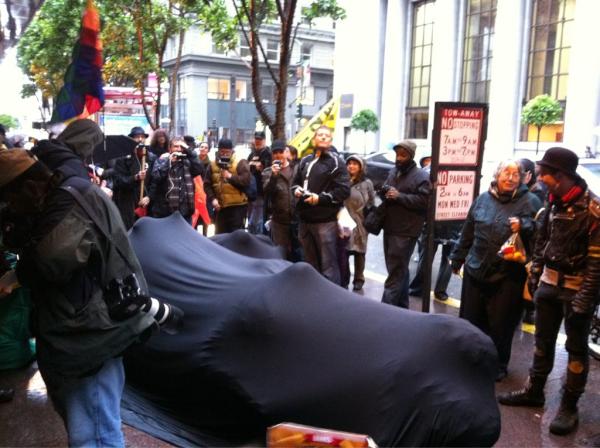 Several people share a single black body sock, seated on the street in San Francisco with an Occupy event. They resisted police arrest for 10 hours.