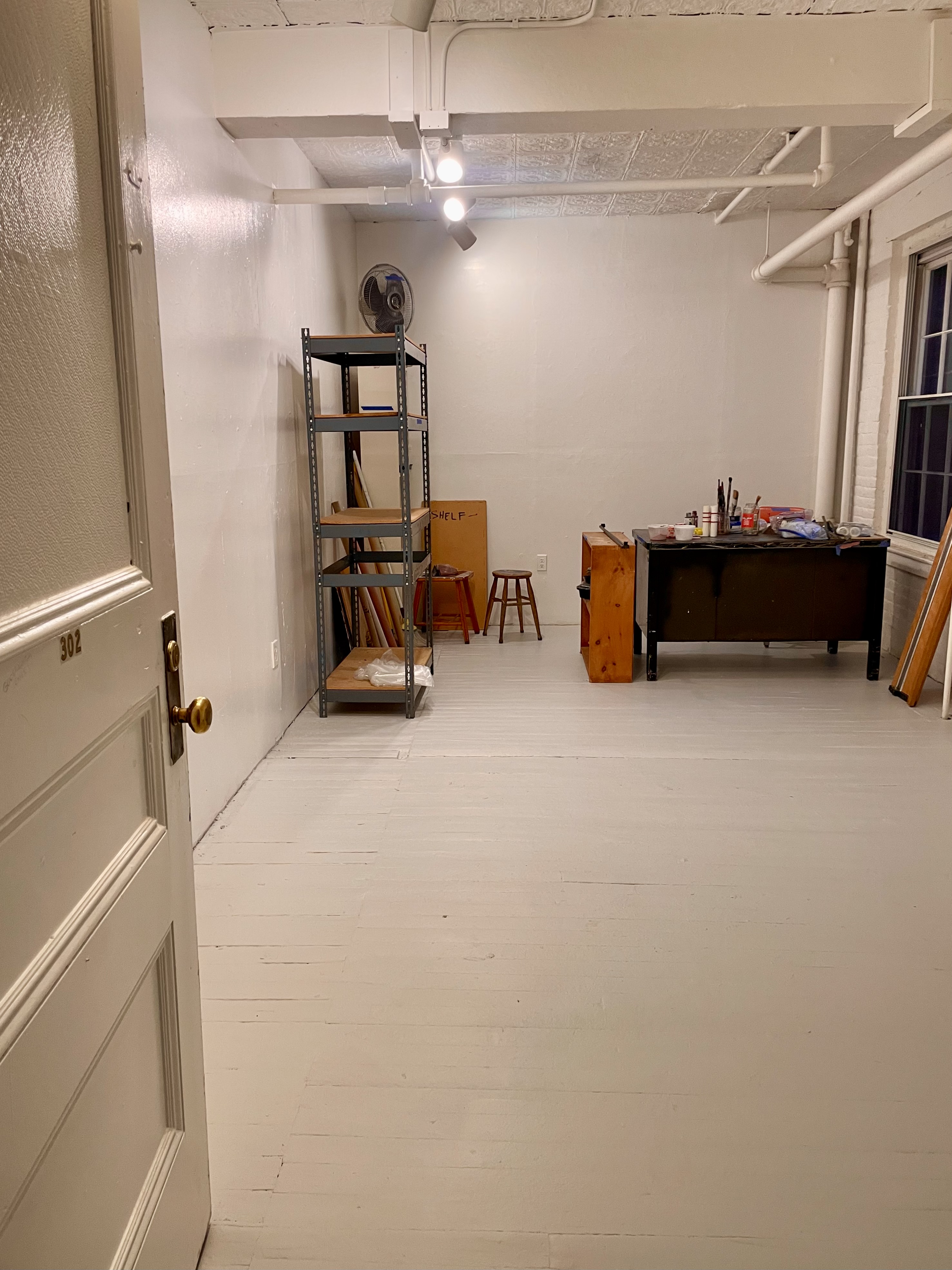 a white walled and white floored studio with desk and shelves awaits new art work.jpeg