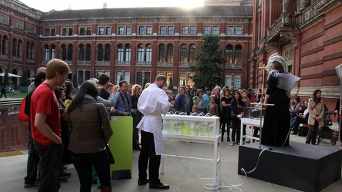 An exterior shot of the V&A museum, where the opera singer performs on stage, flanked by a man in white lab gear poised above a portable set of algae tanks. A crowd listens.