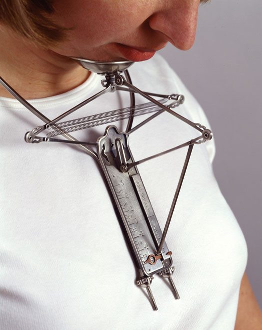 A woman wears a metal "necklace" with a measuring stick attached to a cup for her chin. The vertical measure marks the extroversion level by its distance from the relative elevation of the chin.