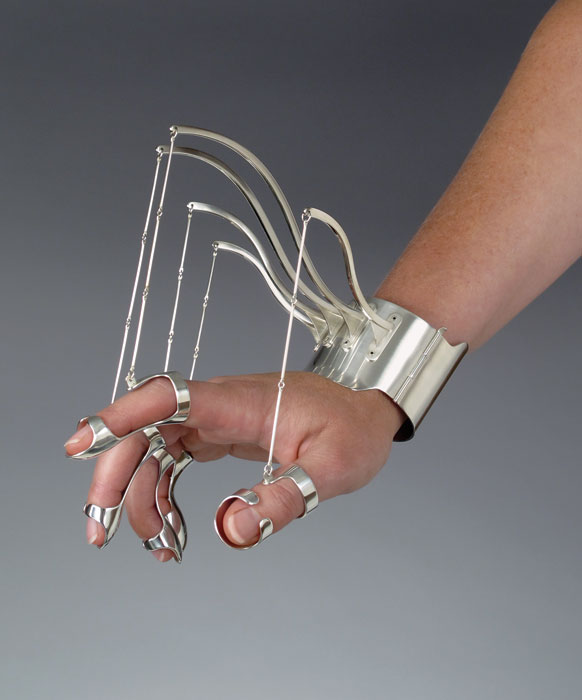 A model wears an aluminum cuff bracelet, from which five metal "marionette" strings hang, with a ring for each finger. When the wearer's hand is braced by these rings and cuff, the just-so arranged hand evokes the poses of classical paintings.