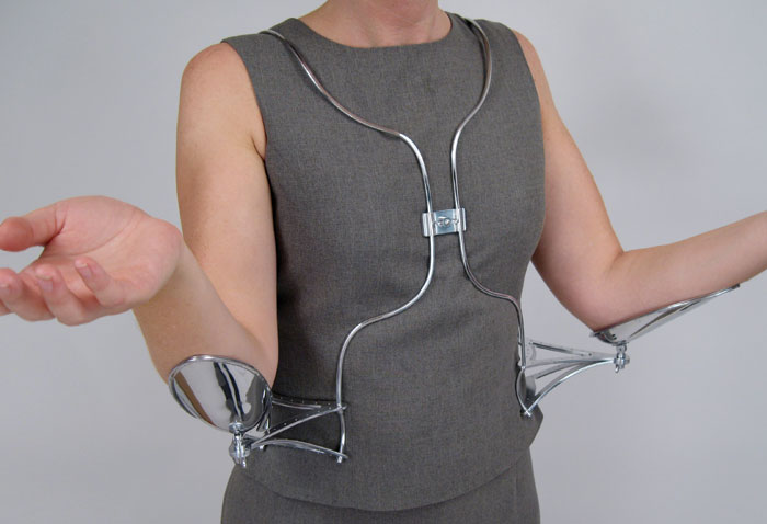 A model wears a metal wire vestment, hung from the neck. Braces mount each side of the wearer's wait, and extensions with cups for the elbows extend to each side. The posture is an open-handed, "unguarded" one.