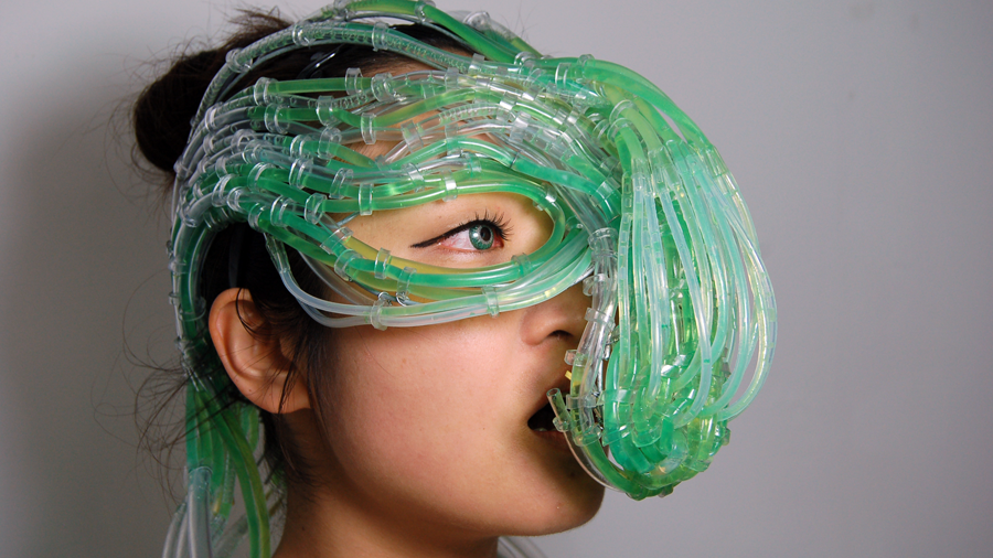 a close up of a woman wearing an elaborate, hydra-like head-and-face wearable of clear plastic tubing, looped and roped together at various intervals, and transporting green alga liquid to the mouth