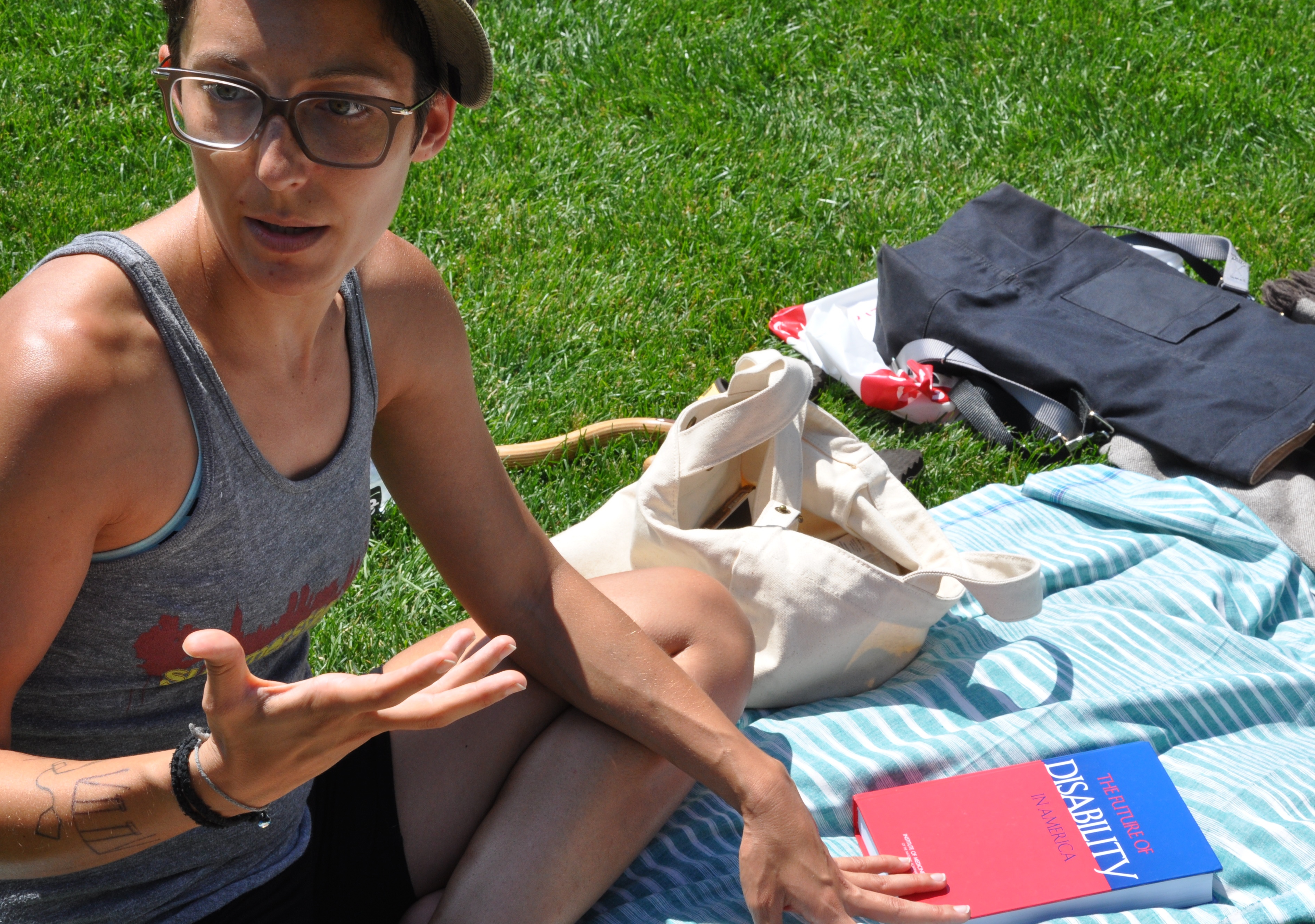 Liz Jackson's bio shot from her site: a woman sitting on a picnic blanket in the sun, her head and one hand engaged mid-conversation with someone off screen, and the other hand resting on a text: The Future of Disability.