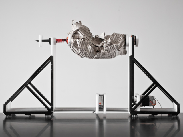 A crumpled piece of newspaper is sandwiched and suspended between two tabletop "vice grip" structures, capable of spinning the object laterally, like a rotisserie. A small squarish tool below the paper scans its surfaces and translates them into sounds.