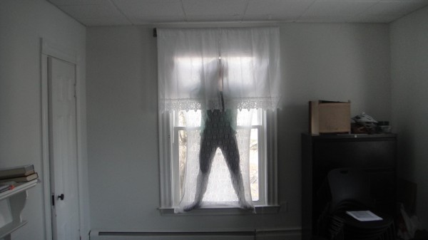 A body sock made from stretchy knitted lace, like so many front window curtains. The wearer steps inside the lace envelop and can disappear, perched in the window frame.