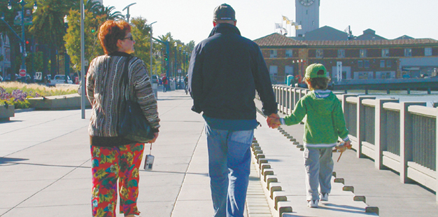 an adult man holds a small child's hand, walking side by side. the child walks on a raised wall or edifice, making their hands' height more equal. 